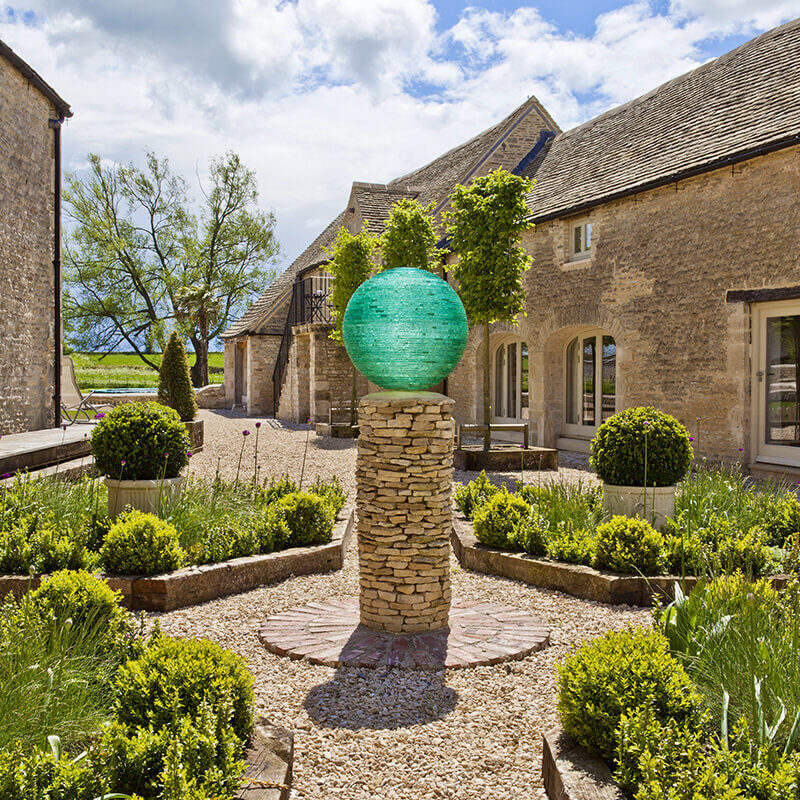 Aurora glass sphere sculpture by James Parker, formed from layered translucent glass and installed on a dry-stone plinth in a garden setting.