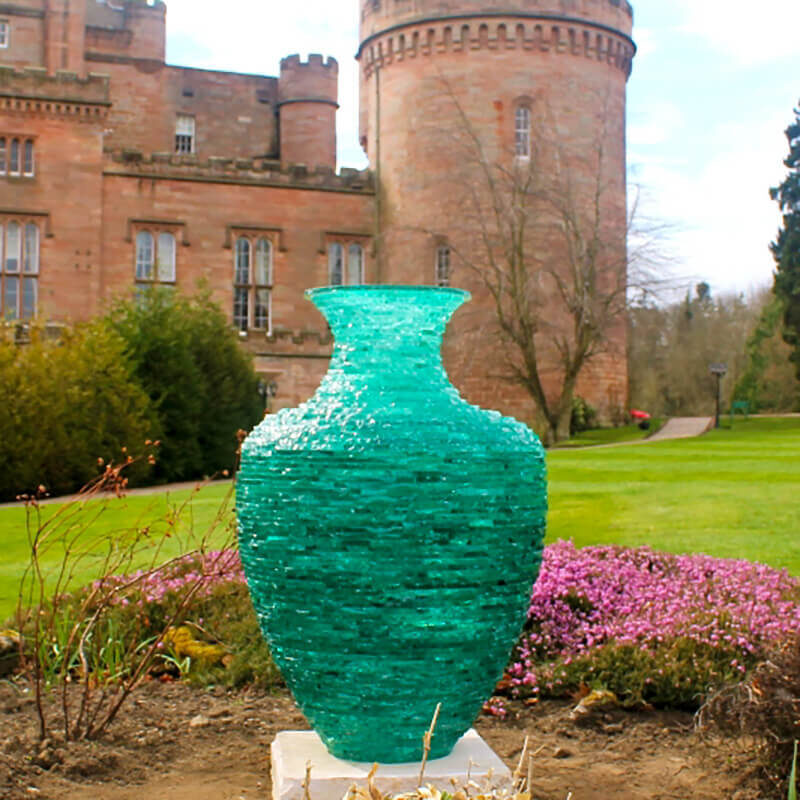 Stacked glass sculpture “Catching the Light” by James Parker, a translucent green Grecian urn form installed outdoors on a sandstone base.
