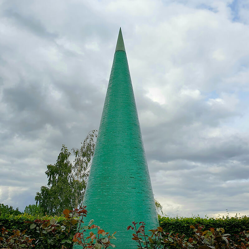 Monumental stacked glass Cone sculpture by James Parker with bronze finial, showing green light refraction through layered glass against the sky.