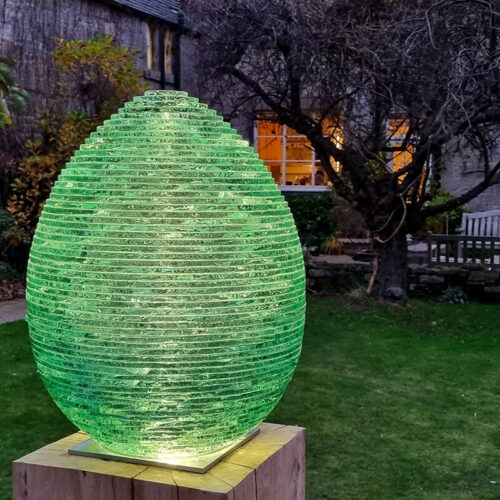 Illuminated stacked glass egg sculpture by James Parker glowing at dusk, lit from within in a landscaped garden.