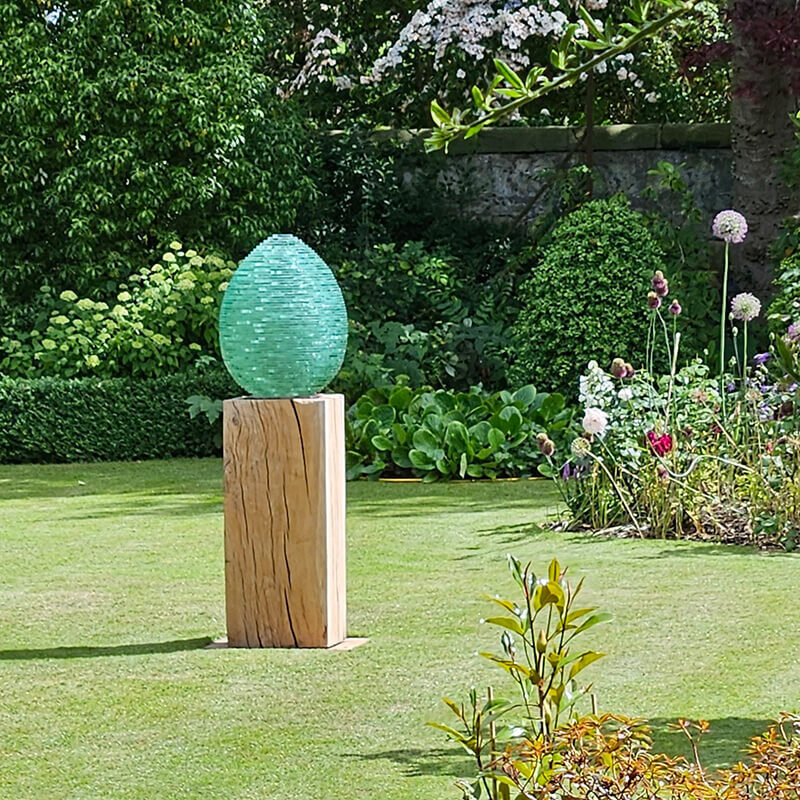 Stacked glass egg sculpture “Creation” by James Parker on a bespoke oak plinth, installed in a private garden setting.