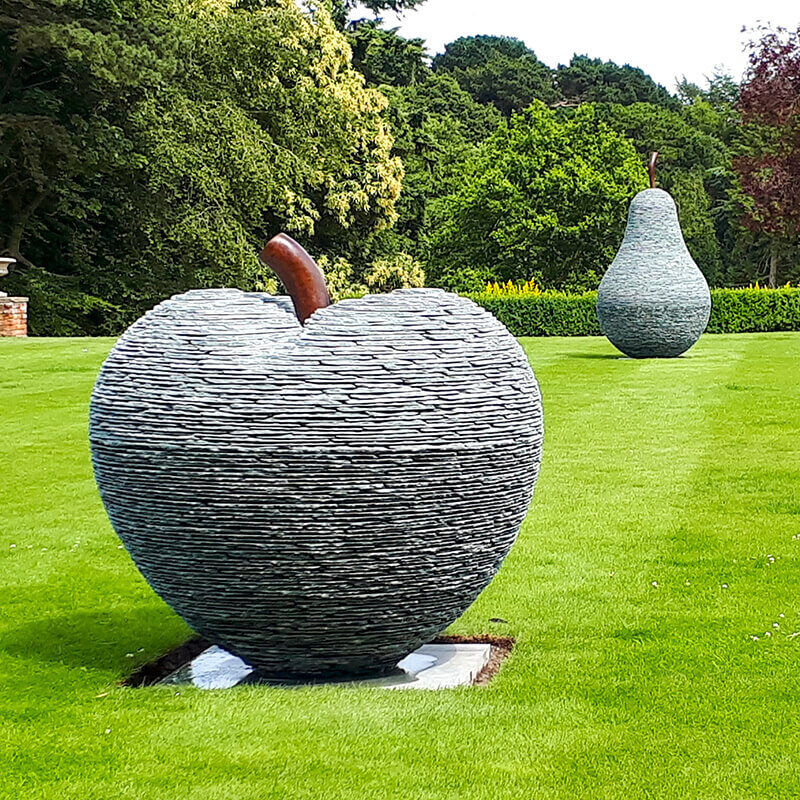 Large stacked slate apple and pear sculptures by James Parker, formed from layered slate with bronze stalks and installed within the grounds of a private country estate.