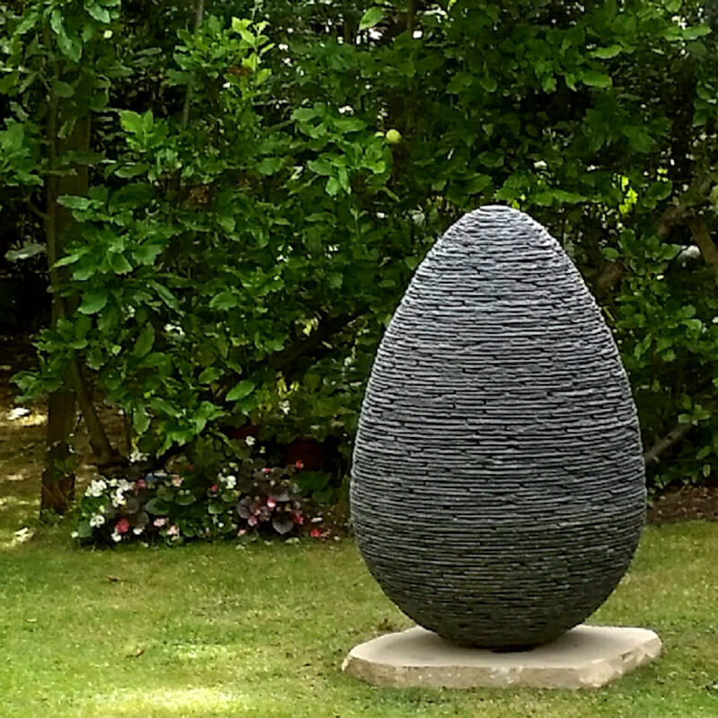 Smaller stacked slate egg sculpture by James Parker in Lakeland Blue slate, positioned within a landscaped garden.