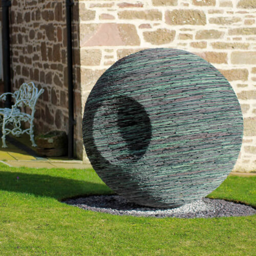 Contemporary slate sphere sculpture “Ever Watchful” by James Parker installed outdoors on a natural boulder base.