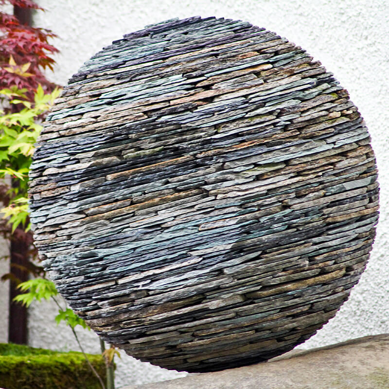 Ever Watchful stacked slate sphere sculpture by James Parker featuring a concave eye form and layered stone construction in a garden setting.