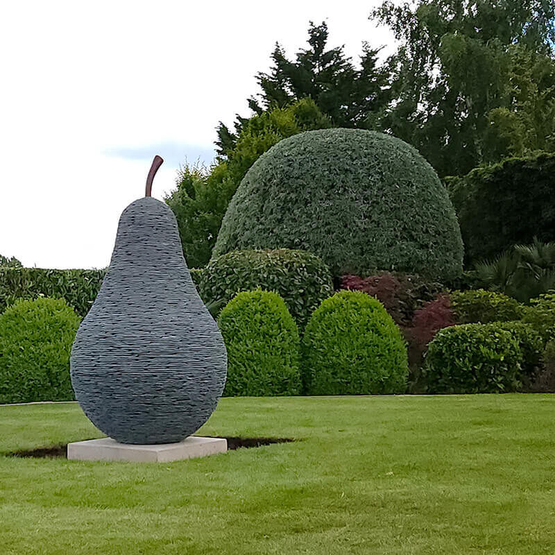 Stacked slate pear sculpture by James Parker with bronze stem, installed as a garden focal point on a sandstone base.