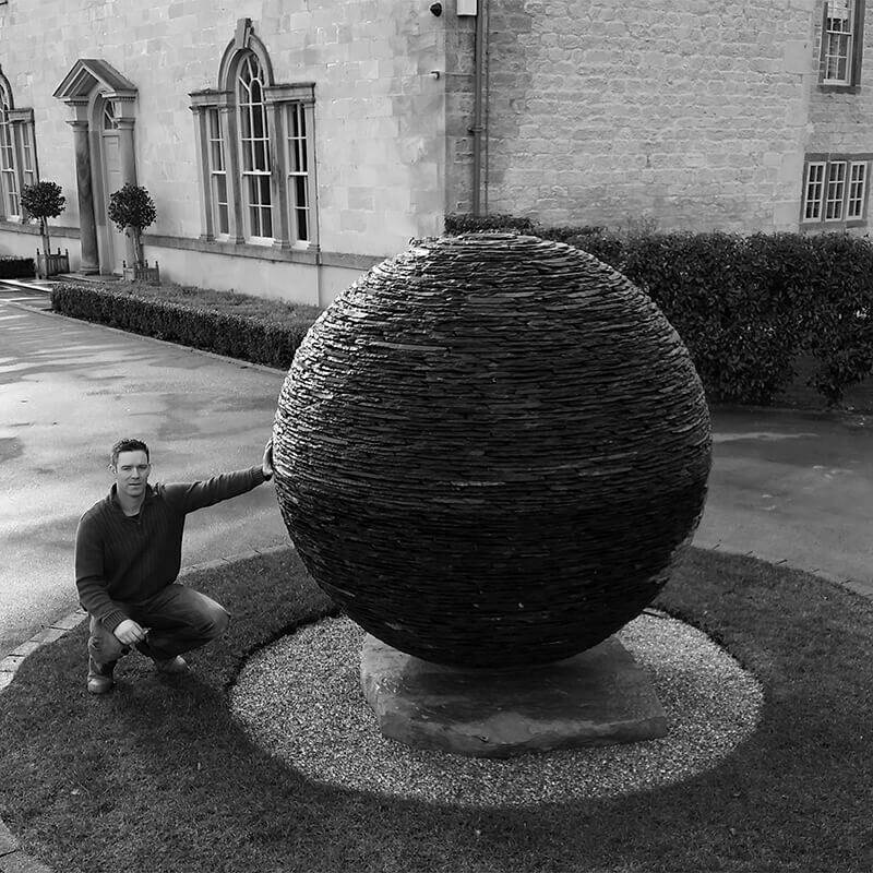 Monumental stacked slate sphere sculpture by James Parker positioned on a sandstone base within a landscaped estate setting.