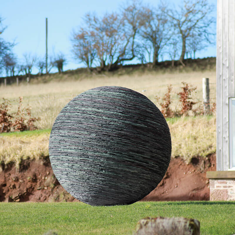 Monumental stacked slate sphere sculpture by James Parker set on a slight tilt within a rural landscape.