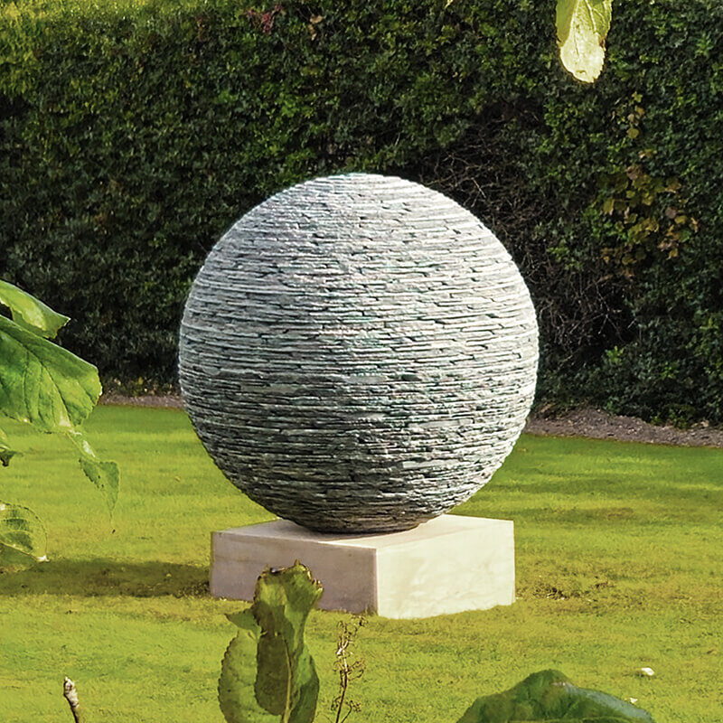 Stacked slate sphere sculpture by James Parker installed on a sandstone plinth in a private garden.