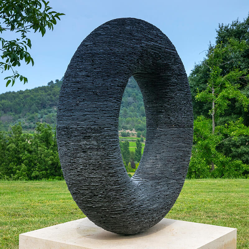 Stacked black slate torus sculpture “Spirit” by James Parker mounted on a whinstone boulder in a landscape setting.