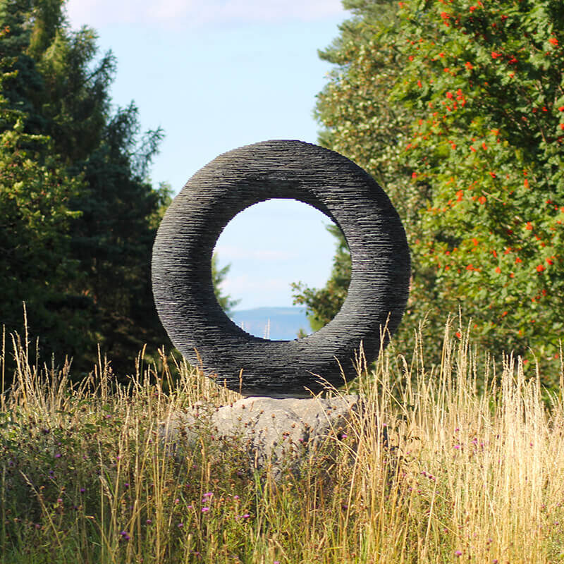 Stacked slate torus sculpture Spirit by James Parker positioned within woodland on a stone base.