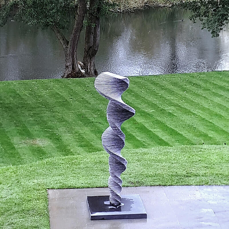 Vortex stacked slate spiral sculpture by James Parker installed beside water in a landscaped garden setting.
