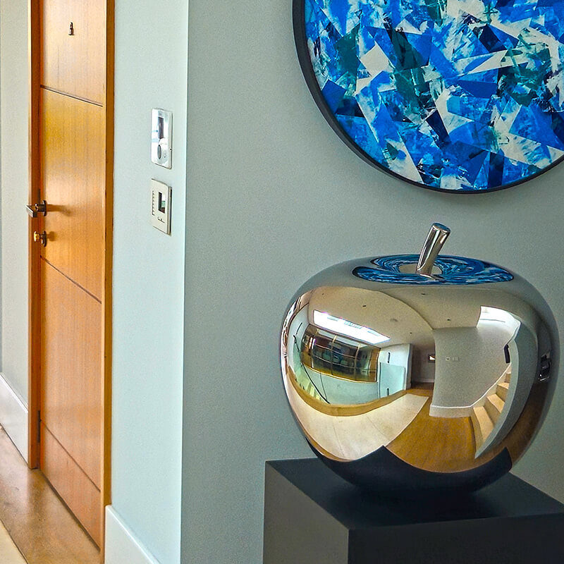 Mirror-polished stainless steel apple sculpture by James Parker displayed on a plinth within a contemporary interior setting.
