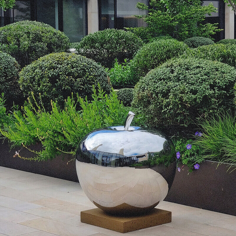 Mirror-polished stainless steel apple sculpture installed on a rooftop terrace with architectural planting.