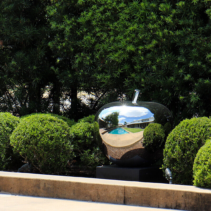 Large mirror-polished stainless steel apple sculpture positioned beside water within a landscaped garden setting.