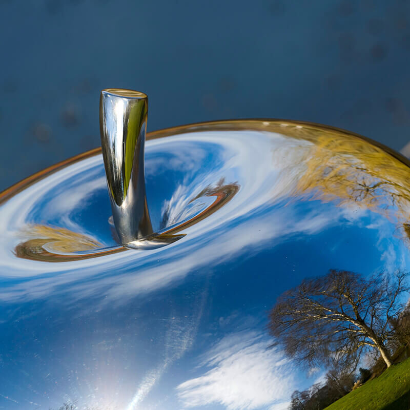 Close-up of mirror-polished stainless steel apple sculpture reflecting sky and surrounding landscape.