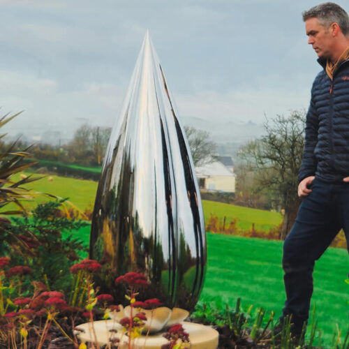 Mirror-polished stainless steel Bud sculpture by James Parker installed on a sandstone base within a landscaped garden setting.