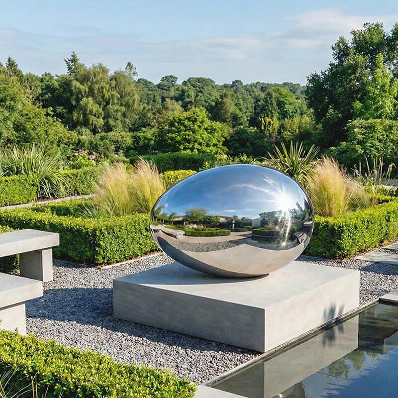 Mirror-polished stainless steel egg sculpture “Creation” by James Parker on a stone plinth beside water in a contemporary garden setting.