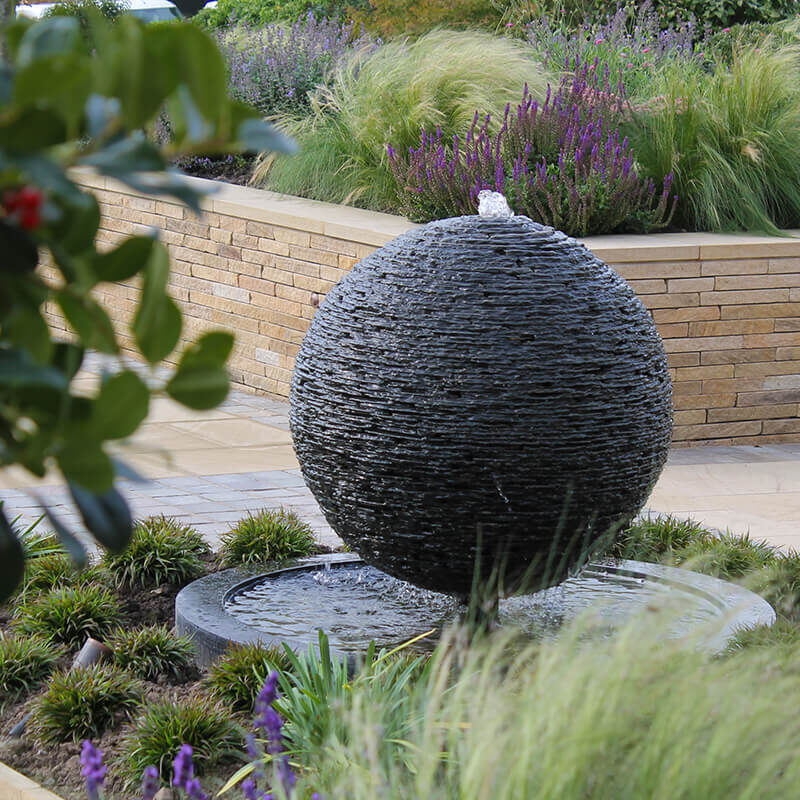 Slate sphere water sculpture by James Parker set within a North Yorkshire garden, water gently cascading over layered stone.