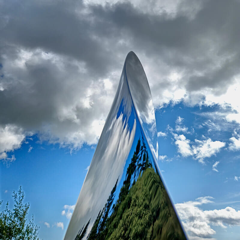 Low-angle view of mirror-polished stainless steel Leaf sculpture reflecting clouds and sky.