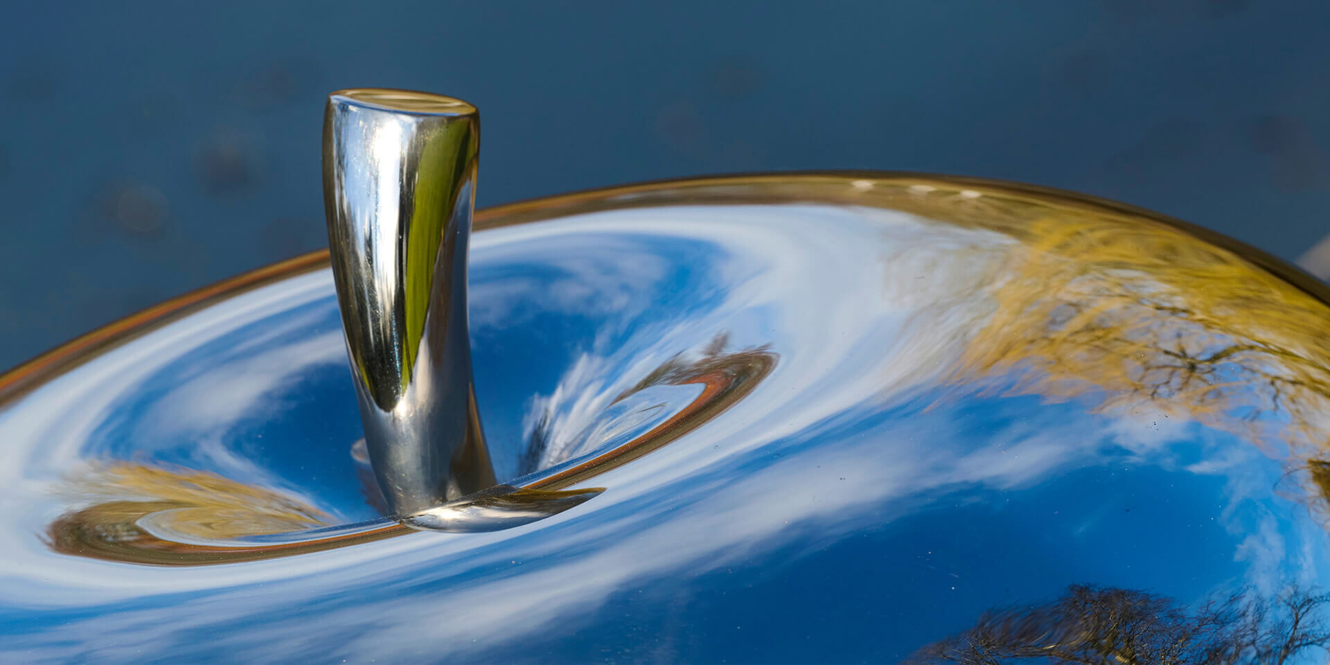 Close-up detail of a mirror-polished stainless steel apple sculpture by James Parker reflecting clouds and sky.