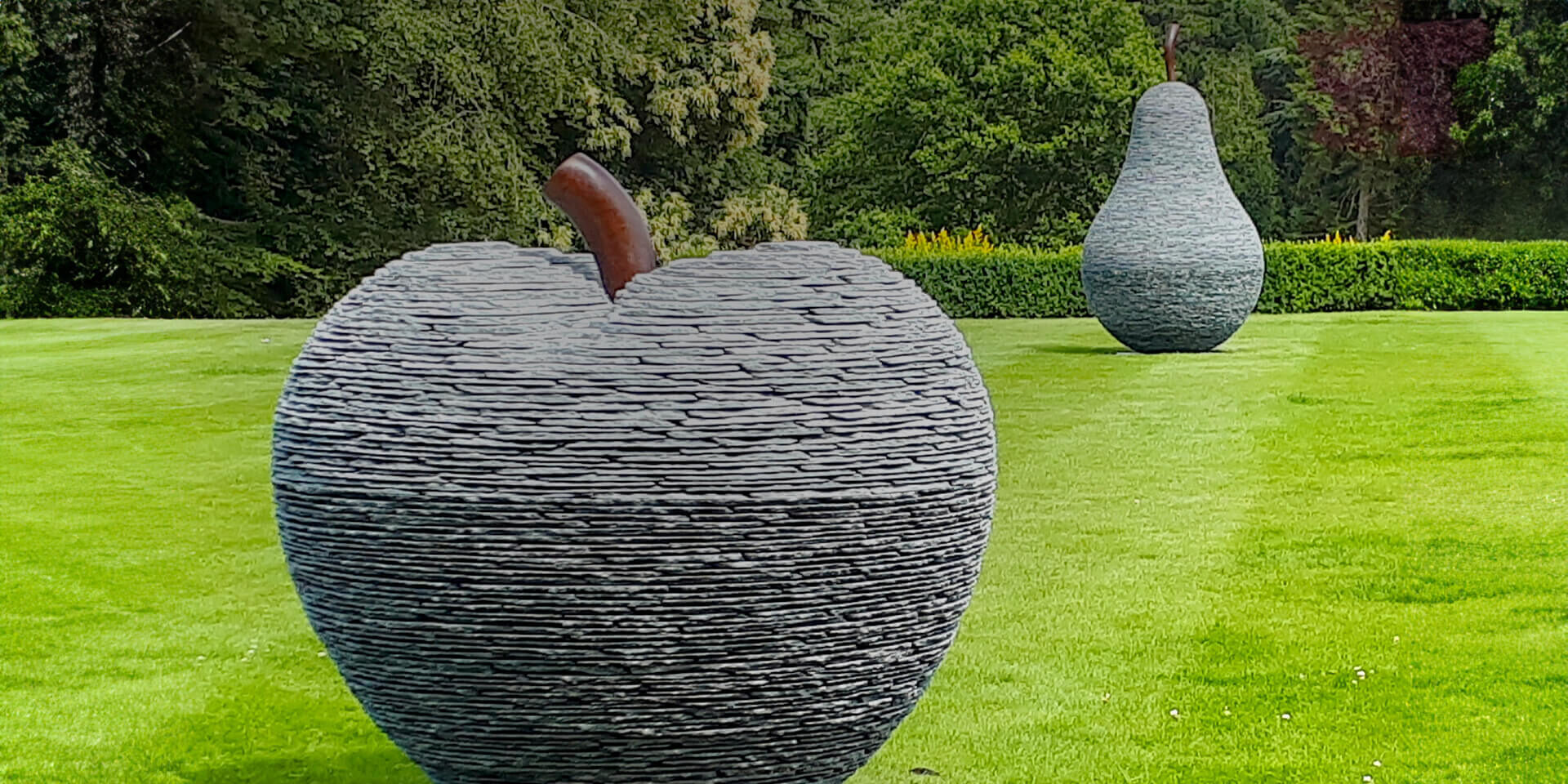 Large stacked slate apple and pear sculptures by James Parker, formed from layered slate with bronze stalks and installed within the grounds of a private country estate.