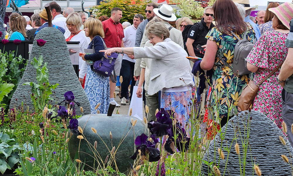 Crowd gathered at James Parker Sculpture trade stand, visitor pointing towards stacked slate and bronze sculptures at RHS Chelsea Flower Show 2023
