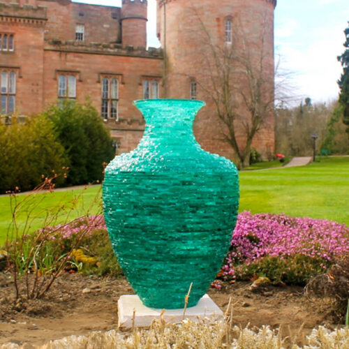 Stacked glass sculpture “Catching the Light” by James Parker, a translucent green Grecian urn form installed outdoors on a sandstone base.