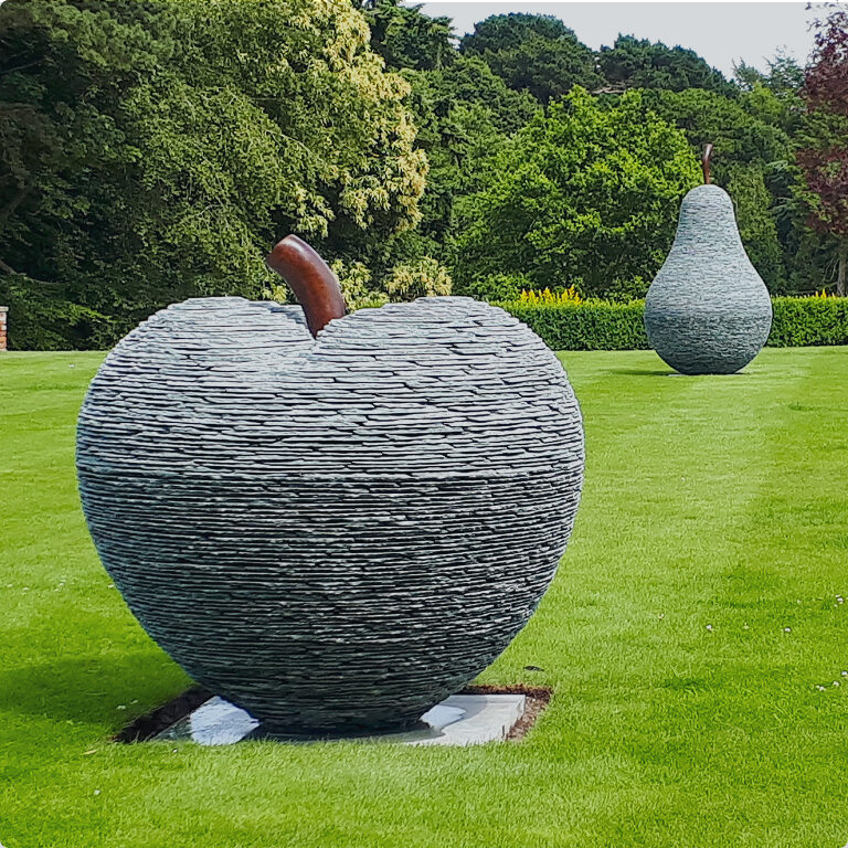 Large stacked slate apple and pear sculptures by James Parker, formed from layered slate with bronze stalks and installed within the grounds of a private country estate.