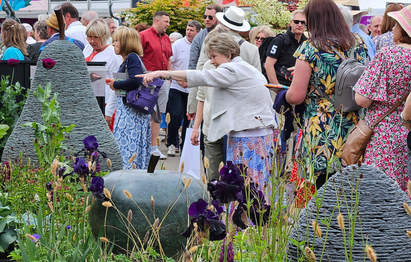 Crowd gathered at James Parker Sculpture trade stand, visitor pointing towards stacked slate and bronze sculptures at RHS Chelsea Flower Show 2023