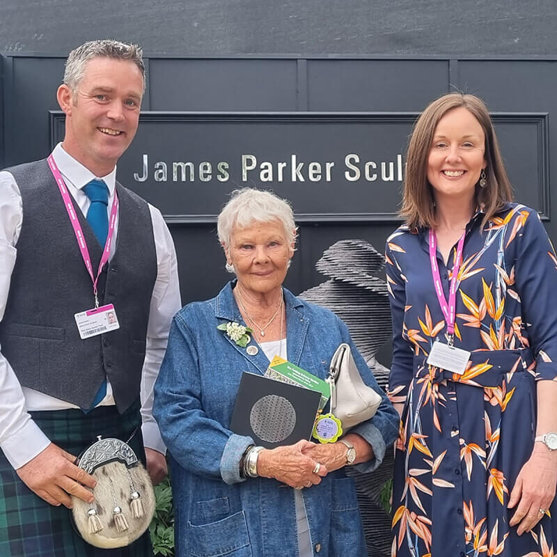 Dame Judi Dench with James and Lesley Parker at the James Parker Sculpture trade stand, RHS Chelsea Flower Show, in front of a slate sculpture display.