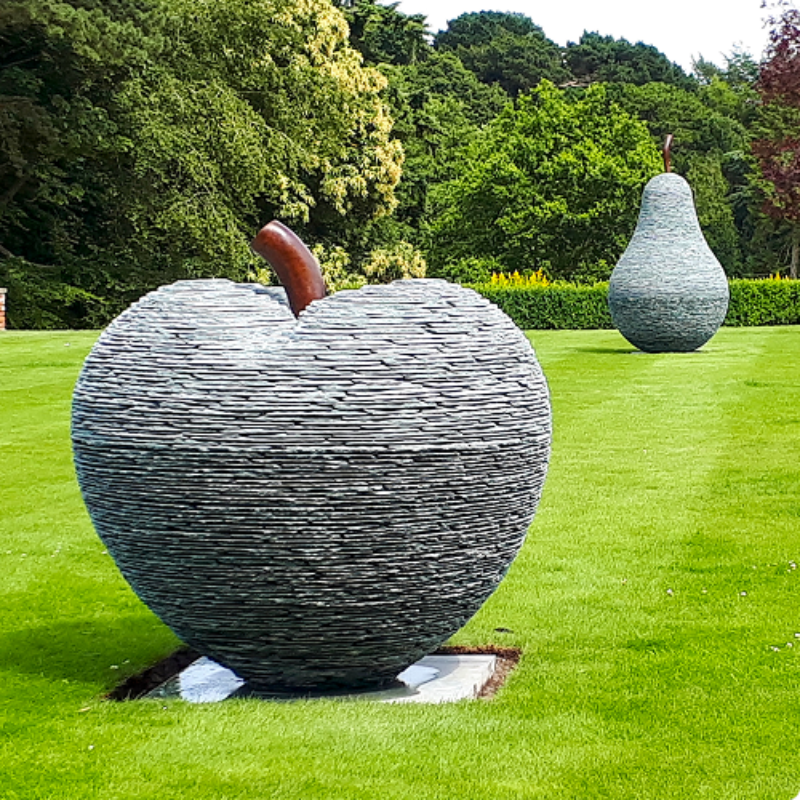 Large stacked slate apple and pear sculptures by James Parker, formed from layered slate with bronze stalks and installed within the grounds of a private country estate.